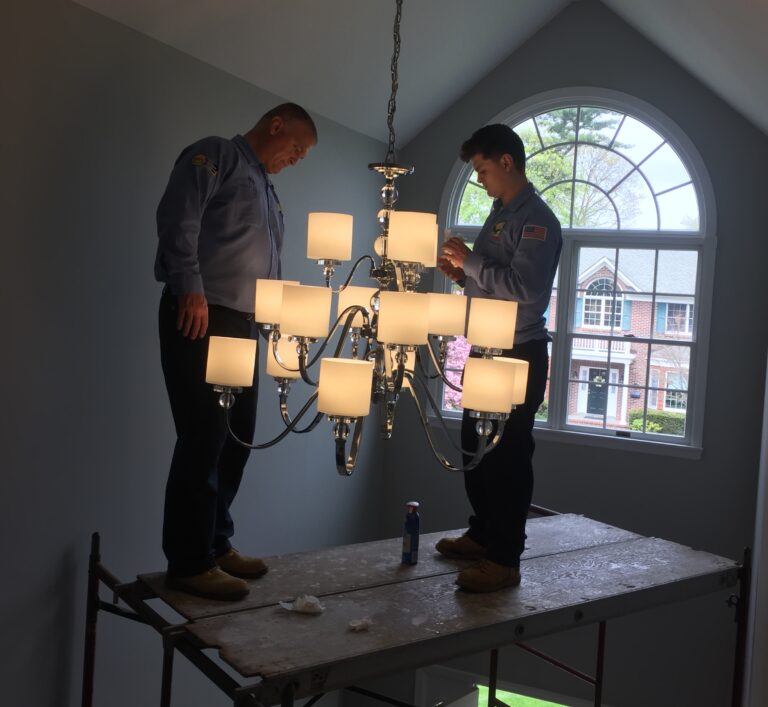 two electricians standing on a platform hanging a chandelier in a home
