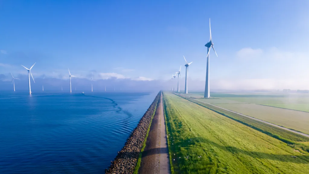 A serene landscape featuring a row of wind turbines along a narrow strip of land between a calm body of water on one side and a green field on the other. The sky is clear with a gentle mist in the distance, and the early morning light casts a tranquil glow over the scene."