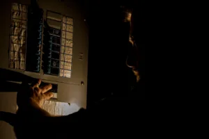 A man, visible only as a faint silhouette, is using a flashlight to investigate a home fuse box in his basement during a power outage.